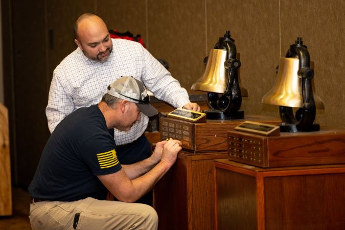 Heartland Division General Manager Tony Fulton watches as Aaron Stich, electrician, applies a nameplate to one of their bells. 